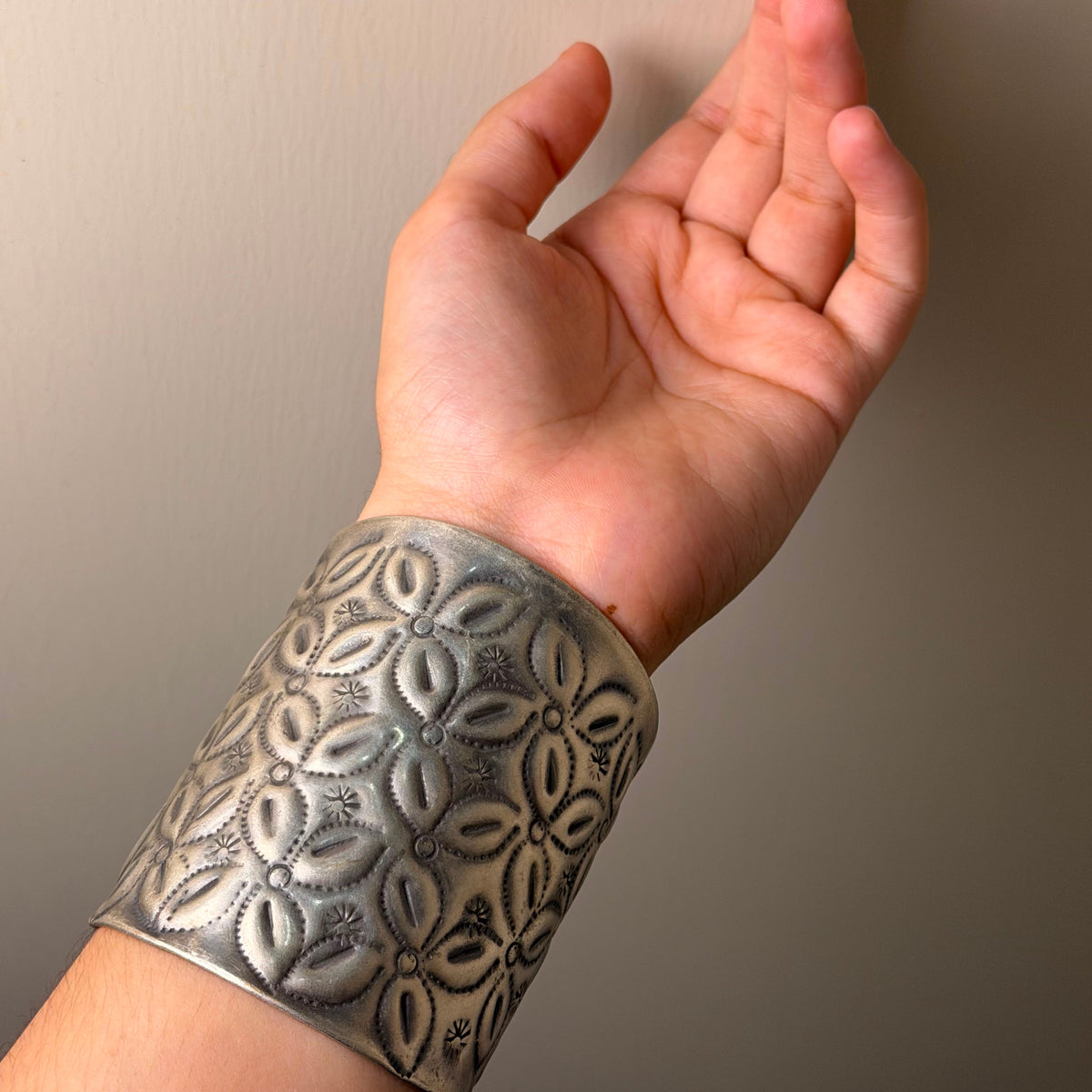 A wide silver cuff bracelet with an embossed floral pattern, worn on a person's wrist against a plain beige wall.
