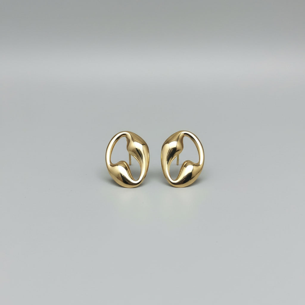 A pair of polished gold-toned abstract loop stud earrings resting on a glossy white ceramic surface, with a bamboo slat background.
