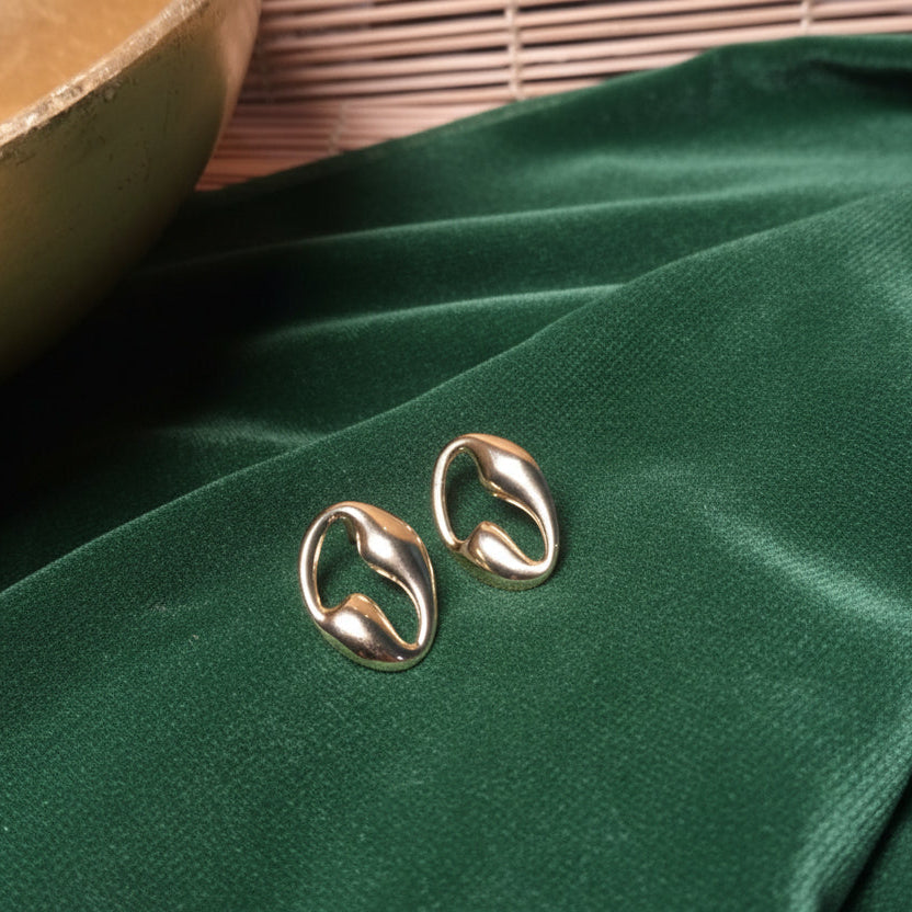 A pair of gold-toned oval hoop earrings resting on draped taupe fabric, with a wooden bowl and bamboo mat in the background.